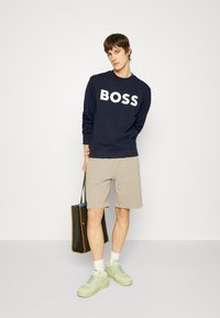 Sweatshirt azul-marinho com grande logotipo branco "BOSS", calções bege claros, ténis verdes e uma mala de tote preta com acentos laranja.