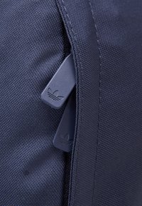 Námorně modrá textilní taška s texturovaným povrchem; obsahuje dva námorně modré zipy s malým logem na táhlech, zdůrazňující funkčnost a design.