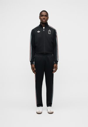 Άνδρας που φορεί μαύρο φόρμα Adidas με λευκές, κόκκινες και μπλε ρίγες κατά μήκος των μανικιών και των ποδιών, στέκεται μπροστά σε απλό φωτεινό φόντο.