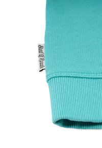 Kangas neulottu turkoosilla, jossa on taitettu hihansuu ja merkkilappu. Materiaalin pinta on sileä ja siinä näkyy tunnistettavia venymiskuvioita.