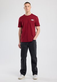 Maroon short-sleeve t-shirt with a logo on the chest, paired with dark jeans and white sneakers on a neutral background.