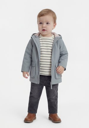 Småbarn iført grå hættetrøje, stribet skjorte, mørke jeans og brune støvler, står foran en ensfarvet hvid baggrund.