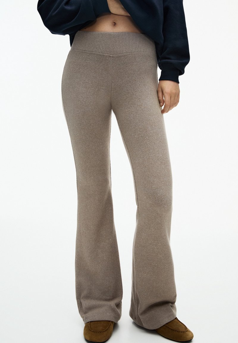 Persoon gekleed in beigekleurige flared high-waist broek, bruine instap schoenen en een donker cropped top, met één hand ontspannen naast zich.