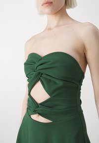 Femme portant une robe bustier vert foncé avec un tissu torsadé et deux découpes à l’avant laissant apparaître la peau.
