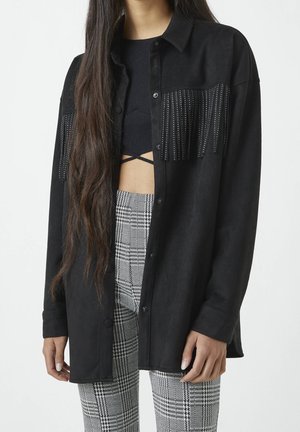 Personne aux cheveux longs portant une chemise noire boutonnée avec des franges cloutées et un crop top noir associé à un pantalon gris à carreaux.