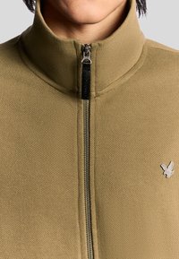 Giacca in felpa con zip di colore verde oliva, con finitura testurizzata. Presenta un colletto alto, zip nera e un accento in argento del logo sul petto.