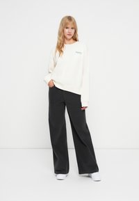 Kremowa bluza z nadrukiem na boku, zestawiona ze spodniami o szerokich nogawkach w kolorze węglowym oraz białymi butami. Gładki materiał, luźny krój i prosty design.