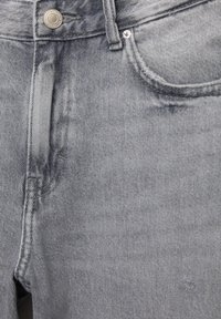 Gros plan sur un jean en denim gris délavé montrant le bouton, la braguette zippée, la poche avant, la passants de ceinture et les détails du tissu texturé.