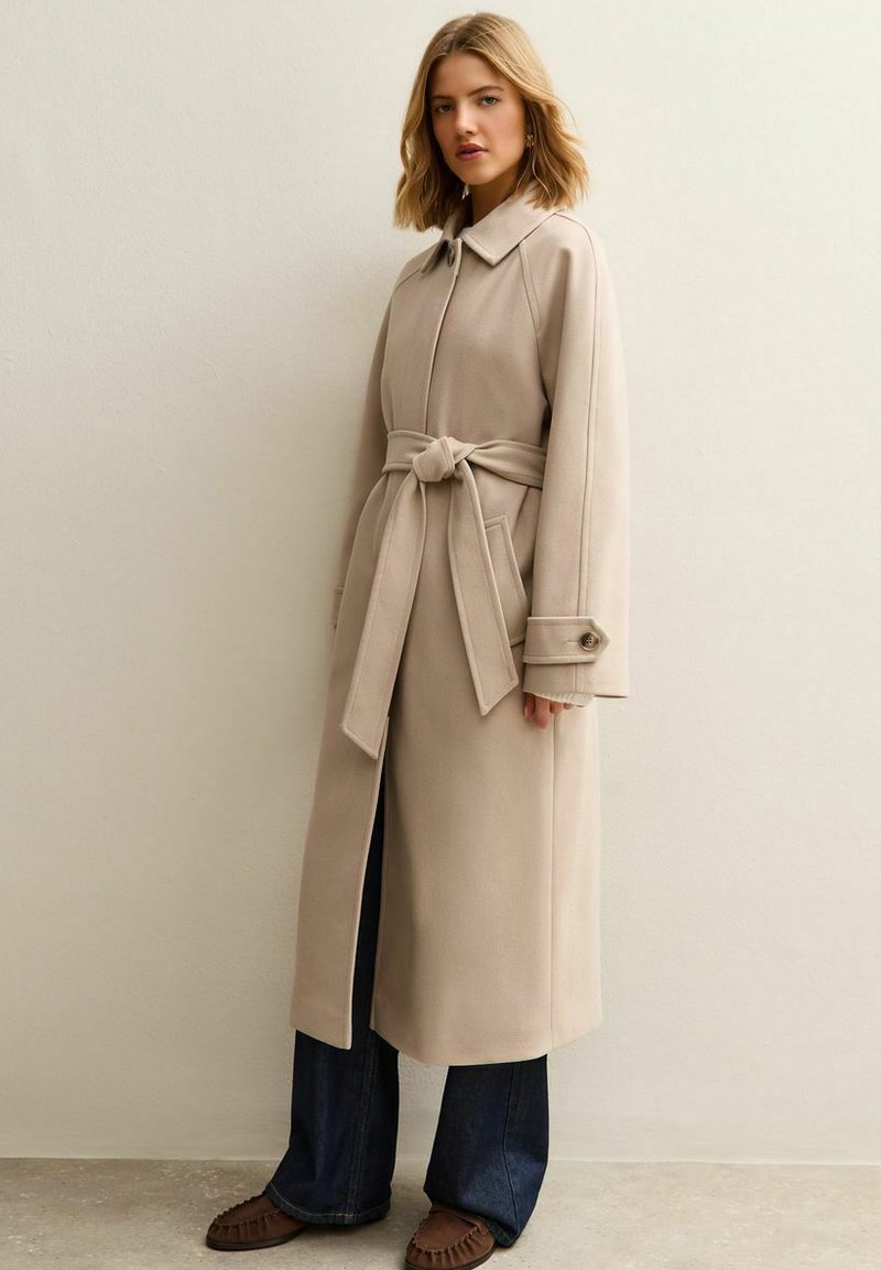 Beżowy płaszcz z paskiem, kołnierzem, oversize'owymi rękawami i dwoma bocznymi kieszeniami, zestawiony z ciemnymi jeansami z rozszerzanymi nogawkami i brązowymi butami.