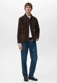 Smeđa jakna od antilopa s ovratnikom, prednjim džepom i kopčama na pritisak, u kombinaciji s plavim trapericama i crnim kožnim cipelama.