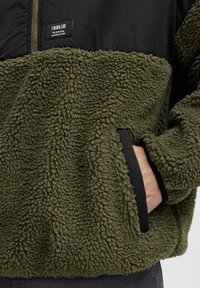 Fleece-pullover i olivgrön, med övre del i svart nylon, strukturerad yta, sidofickor och en varumärkesetikett på bröstet.