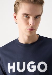 Tricou din bumbac de culoare bleumarin, cu un mare imprimat alb "HUGO" pe față. Guler rotund și mâneci scurte, textură netedă, cu detalii minime.
