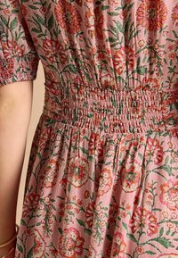 Blumenkleid mit gesmokter Taille. Hat einen rosa Grundton mit roten und grünen Blumen. Kurze Ärmel und fließender Rock aus strukturiertem Stoff.