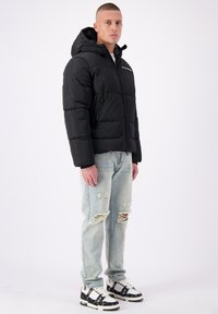 Zwarte pufferjack met een capuchon, voorzien van ritszakken en een gestikt patroon. Gepaard met lichte, versleten jeans en zwarte sneakers.