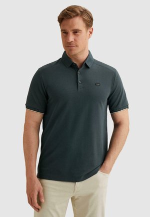 Mann trägt ein dunkelgrünes Poloshirt mit kurzen Ärmeln und hellbeige Hose, steht mit einer Hand in der Tasche und schaut leicht zur Seite.