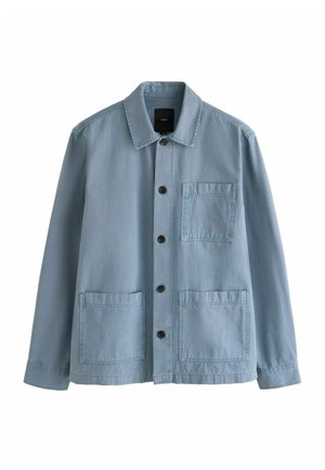 Ung mand iført solbriller, denimjakke, stribet slips, jeans og sorte sko går på en træbevokset grusstien om dagen.