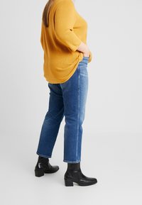 Żółty sweter z prążkowanego materiału, niebieskie dżinsy o lekko zwężanym kroju oraz czarne botki na obcasie z teksturowanym wzorem.