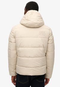 Beige pufferjakke med vandret quiltet mønster, hætte og broderet logo på ryggen. Glat, let materiale, afslappet pasform.