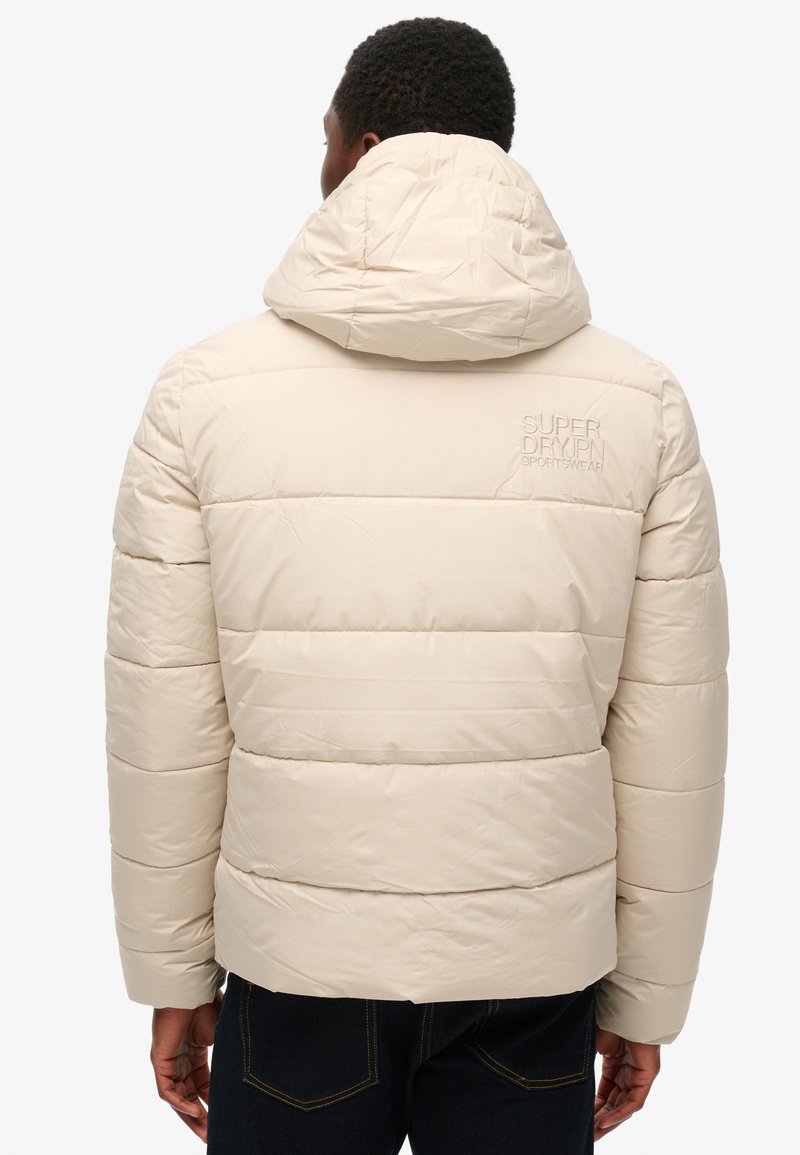 Beige Steppjacke mit horizontalem Steppmuster, Kapuze und gesticktem Logo auf dem Rücken. Glattes, leichtes Material, lässige Passform.