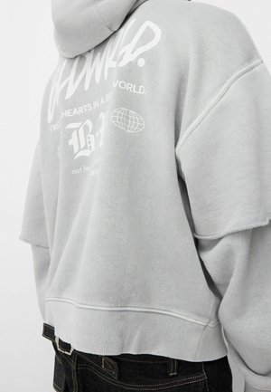 Henkilö, jolla on päällään vaaleanharmaa huppari, jossa on valkoinen teksti ja maapallon kuva takana, yhdistettynä tummiin farkkuihin.