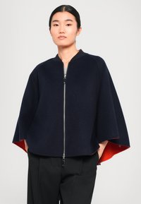 Cape en laine bleu marine avec une fermeture éclair à l'avant, des manches raglan et une doublure rouge vif. Coupe ajustée avec une forme évasée. Tissu texturé.
