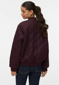 Veste bomber matelassée bordeaux avec poignets et ourlet côtelés. Caractérisée par un col arrondi et un motif en losanges texturé. Portée avec un jean foncé.