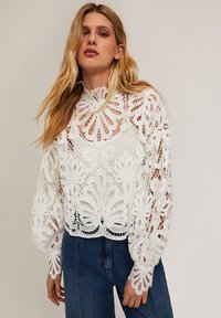 Witte kanten blouse met lange, opgeblazen mouwen. Kenmerkend voor ingewikkelde bloempatronen en een hoge halslijn. Gepaard met blauwe denim jeans.