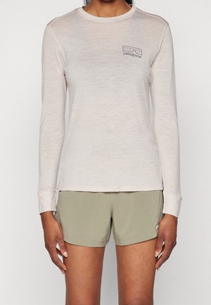 Femme portant une chemise Patagonia beige clair à manches longues et un short vert olive, debout devant un fond blanc uni.