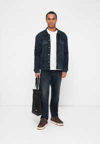 Muškarac u tamnoplavom trapera jakni i trapericama, u bijelom majici, drži crnu torbu, stoji uz bijelu pozadinu.