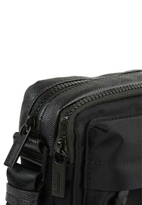 Bolsa de nylon negra con doble cremallera, detalles texturizados, correa ajustable y un acabado suave. Forma rectangular compacta con costuras visibles.