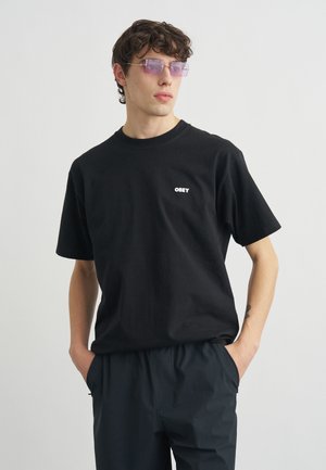 Giovane uomo che indossa occhiali da sole con lenti viola e una maglietta nera con il logo bianco "OBEY", in piedi con le mani in tasca.