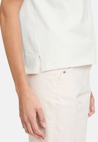 Haut léger en coton blanc avec fentes latérales, présentant des lignes épurées et une coupe décontractée. Associé à un pantalon beige clair et à des accessoires minimalistes.