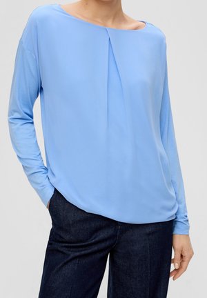 Mujer con blusa de manga larga azul claro con un pliegue central y pantalones azul oscuro, posando con una mano en el bolsillo.