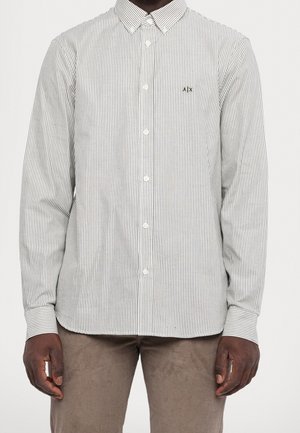 Homme portant une chemise blanche à manches longues avec de fines rayures verticales grises et un pantalon marron, debout les bras détendus.