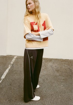Jeune femme aux longs cheveux blonds porte une chemise beige oversize avec un imprimé rouge, manches longues à rayures, pantalons larges noirs et chaussures blanches, debout en plein air.