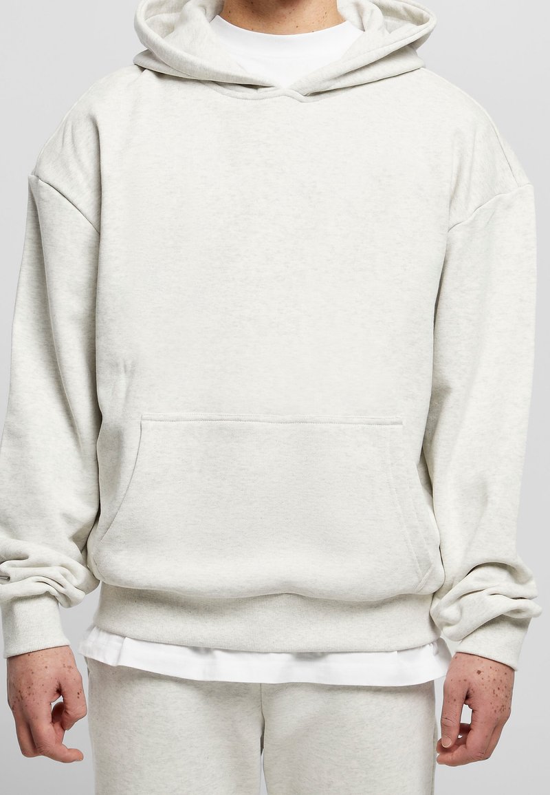 Persona che indossa un ampio hoodie beige chiaro con tasca a marsupio, sovrapposto a una camicia bianca, abbinato a pantaloni della tuta coordinati.