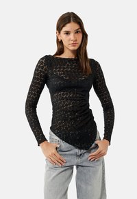Zwarte kanten langslavige top met een aansluitend ontwerp, gestructureerd bloemmotief en een puntige zoom, gecombineerd met lichtgrijze high-waisted jeans.