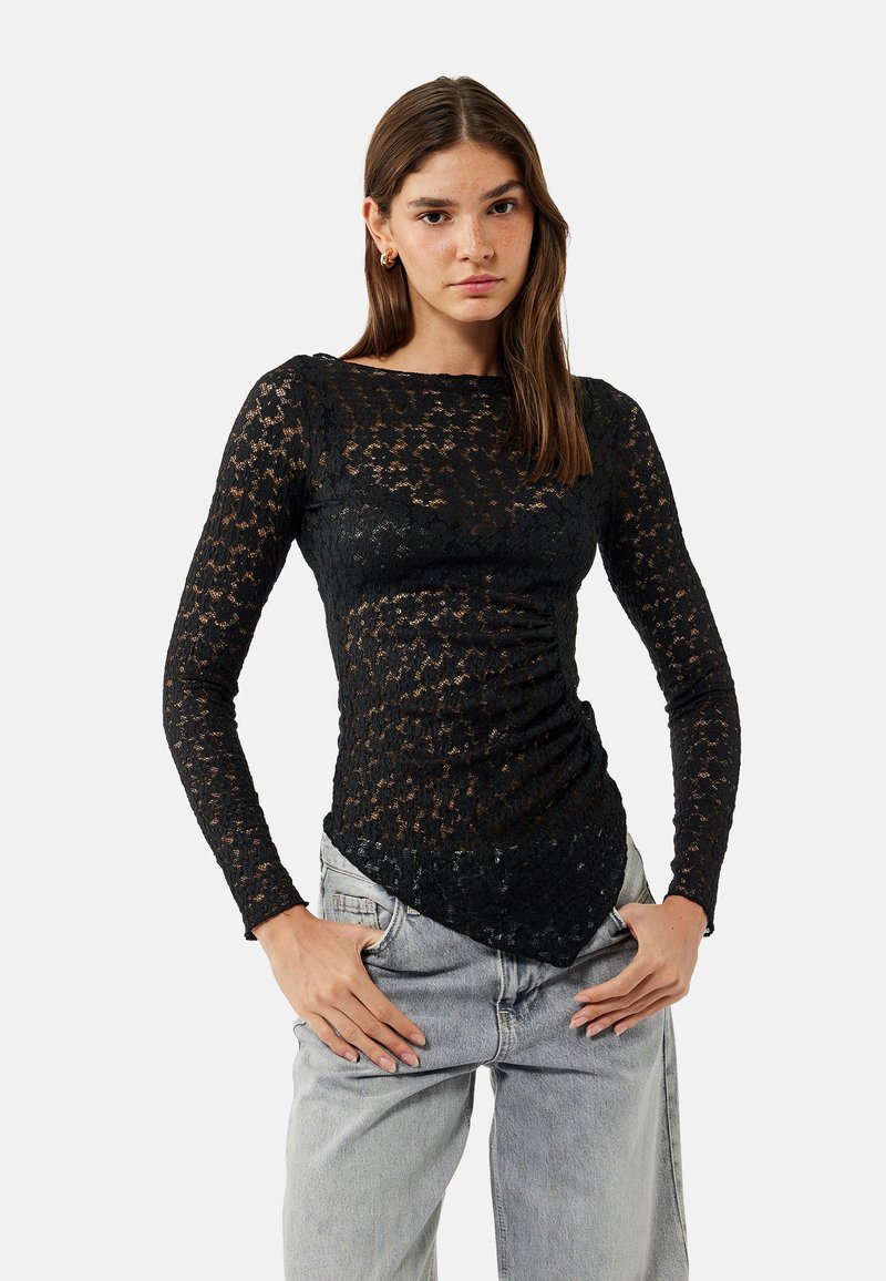 Schwarzes Spitzenshirt mit langen Ärmeln, enganliegendem Design, strukturiertem Blumenmuster und spitzem Saum, kombiniert mit eher grauen High-Waist-Jeans.