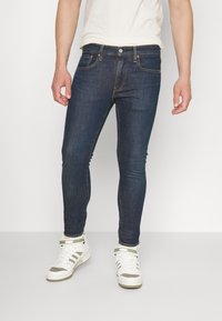 Jeans de denim skinny fit, azul escuro, com design de cinco bolsos, costura clara e fecho de botão, usados com ténis brancos de cano alto.