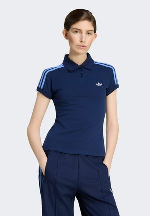 Kvinde med kort brunt hår iført en marineblå Adidas polo skjorte med lyseblå striber og matchende marineblå bukser, hænderne i lommerne.