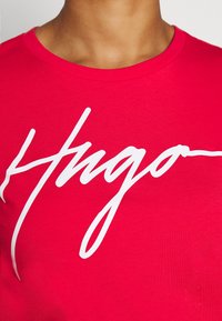 Camiseta de algodón roja con cuello redondo, que presenta el texto en cursiva blanca "Hugo" en el pecho. Textura suave, diseño ajustado.