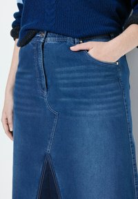 Falda de mezclilla en azul oscuro, con una abertura delantera y costuras en contraste. Incluye un bolsillo y se combina con un suéter de punto texturizado.