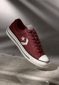 Red leather sneaker with white star logo, textured surface, round toe cap, and white midsole featuring black accent stripes and laces.