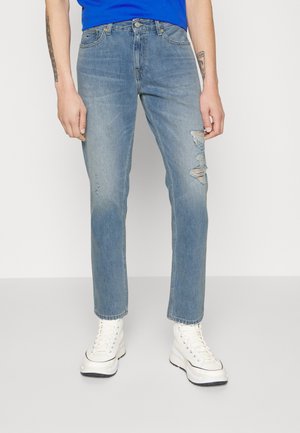 Osoba ubrana w jasnoniebieskie, postarzane dżinsy, białe trampki typu high-top oraz niebieską koszulę, stojąca na tle jednolitego białego tła.