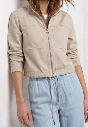 Person trägt eine beige Zip-Up-Jacke mit zwei Brusttaschen und hellblaue Kordelzug-Hose, steht vor einer weißen strukturierten Wand.