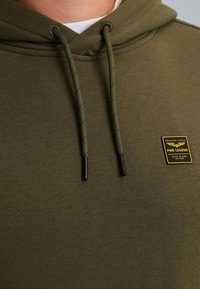 Primo piano di una felpa verde oliva con coulisse e un piccolo patch del logo PME Legend giallo e nero sul petto.