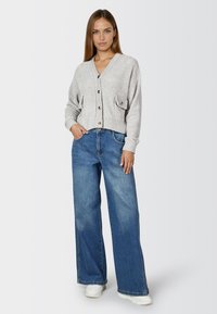 Grau gestrickte Strickjacke mit V-Ausschnitt, Fronttaschen und Knöpfen, kombiniert mit weit geschnittenen blauen Jeans und weißen Sneakern.