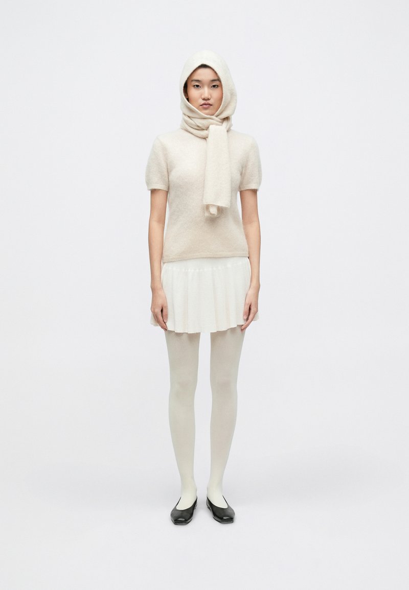 Bluză din lână deschisă la culoare, cu mâneci scurte, asortată cu o fustă mini plisată, ciorapi opaci și balerini negri. Modelul poartă o eșarfă asortată.