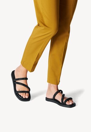 Sandales noires avec plusieurs lanières scintillantes, semelle plate et design ouvert à l'avant, associées à un pantalon tapered de couleur moutarde.