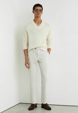 Mann trägt cremefarbenen V-Ausschnitt-Pullover, hellbeige Hose, dunkelbraune Loafers und Sonnenbrille, steht mit den Händen in den Taschen vor einer weißen Wand.